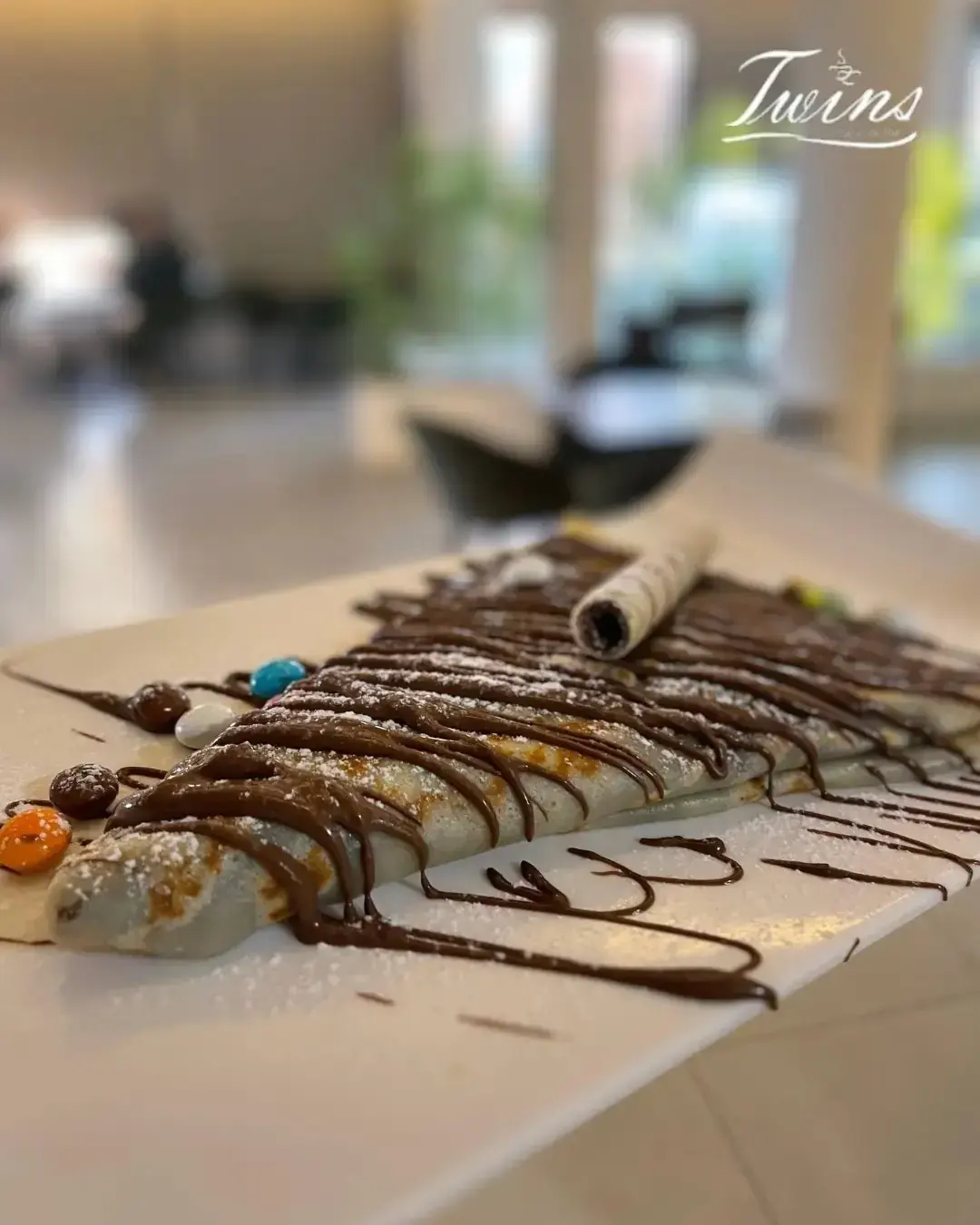 Crêpe nappée de chocolat fondu, sucre glace et bonbons colorés, sur une assiette rectangulaire.