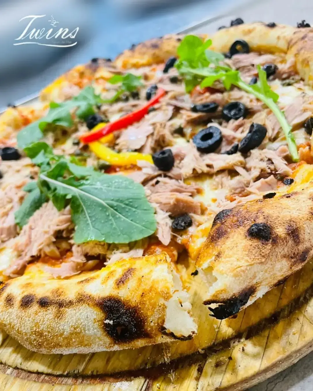 Pizza au thon, olives noires, poivrons et roquette, servie sur une planche en bois.