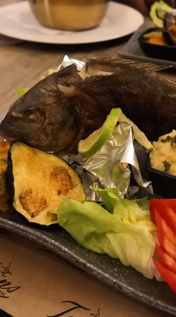 Un poisson entier grillé, servi avec aubergine grillée, citron vert, salade, tomate et purée.