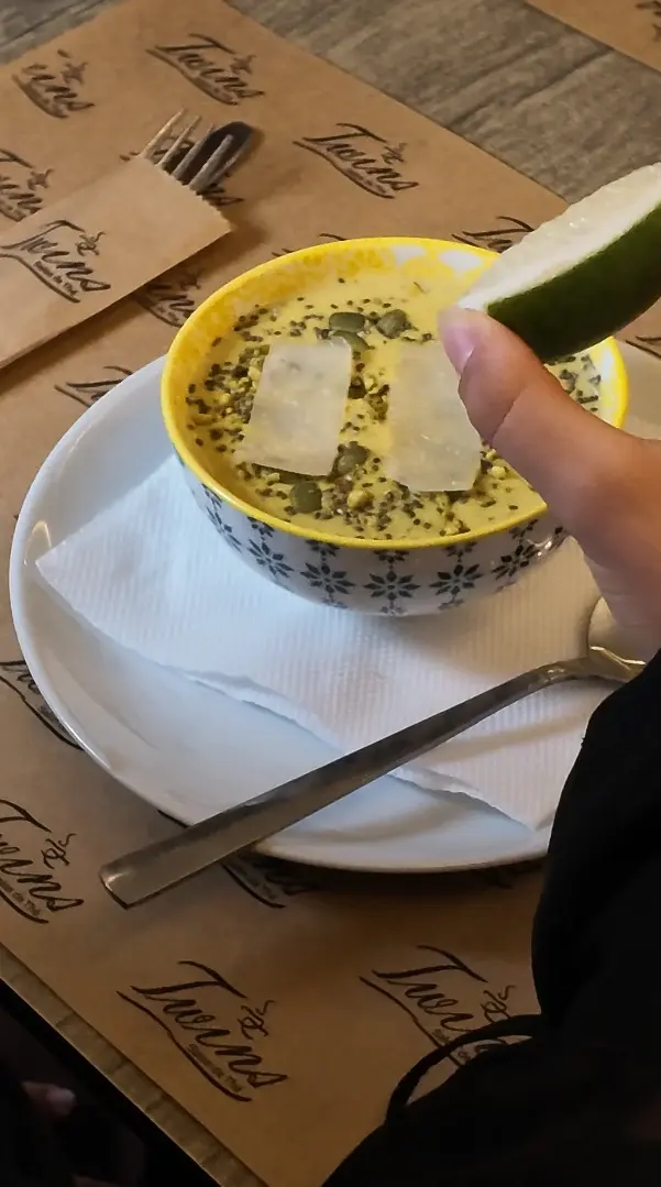 Un bol de soupe veloutée parsemée de graines et de copeaux de fromage, avec un quartier de citron vert.
