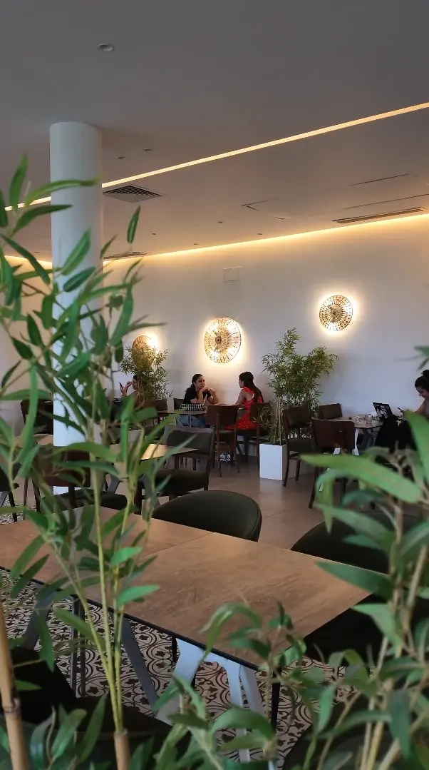 La salle du Twins derrière un rideau de feuillage : tables en bois, chaises vertes et appliques rondes dorées.