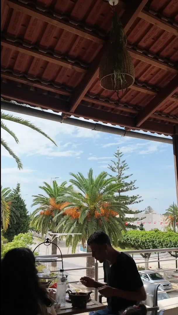 Sous la pergola en bois de la terrasse, des clients déjeunent face aux palmiers et au ciel bleu.