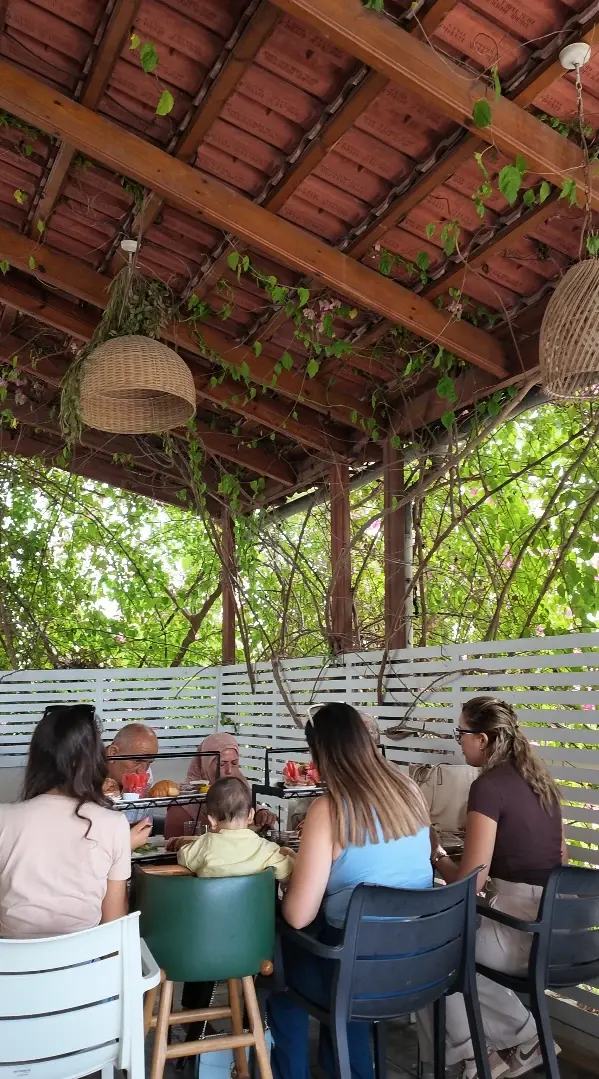 Une famille attablée sous la pergola en bois de la terrasse, couverte de plantes grimpantes et de suspensions en osier.