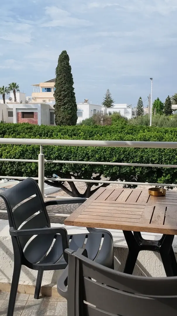 Une table en bois de la terrasse, deux chaises et la vue sur les haies, un cyprès et les maisons blanches.